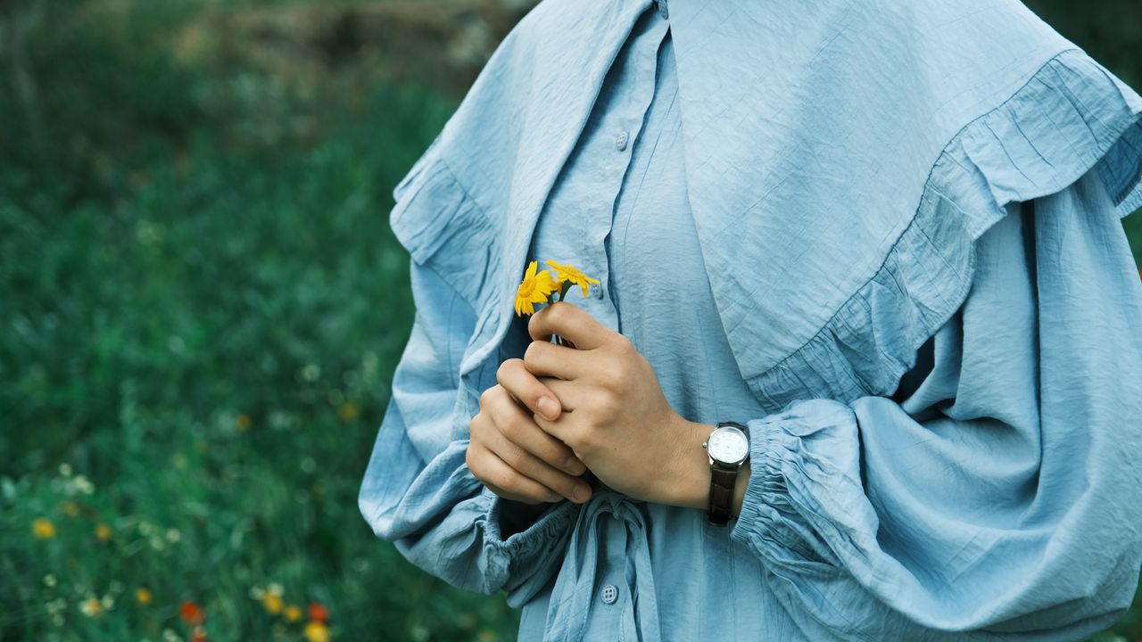 A woman in a blue dress holds a yellow flower in a green outdoor setting, capturing a serene moment.
